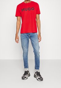 Camiseta de algodón roja con "HUGO" en letras negras, combinada con jeans de mezclilla azul y zapatillas negras y blancas. Ajuste simple y casual.