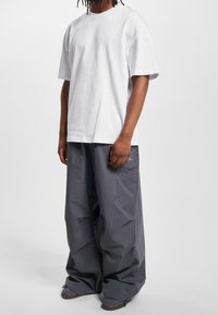 Bílý bavlněný tričko s volným střihem, doplněné volnými tmavě šedými kalhotami. Kalhoty mají nenápadné logo poblíž pasu.