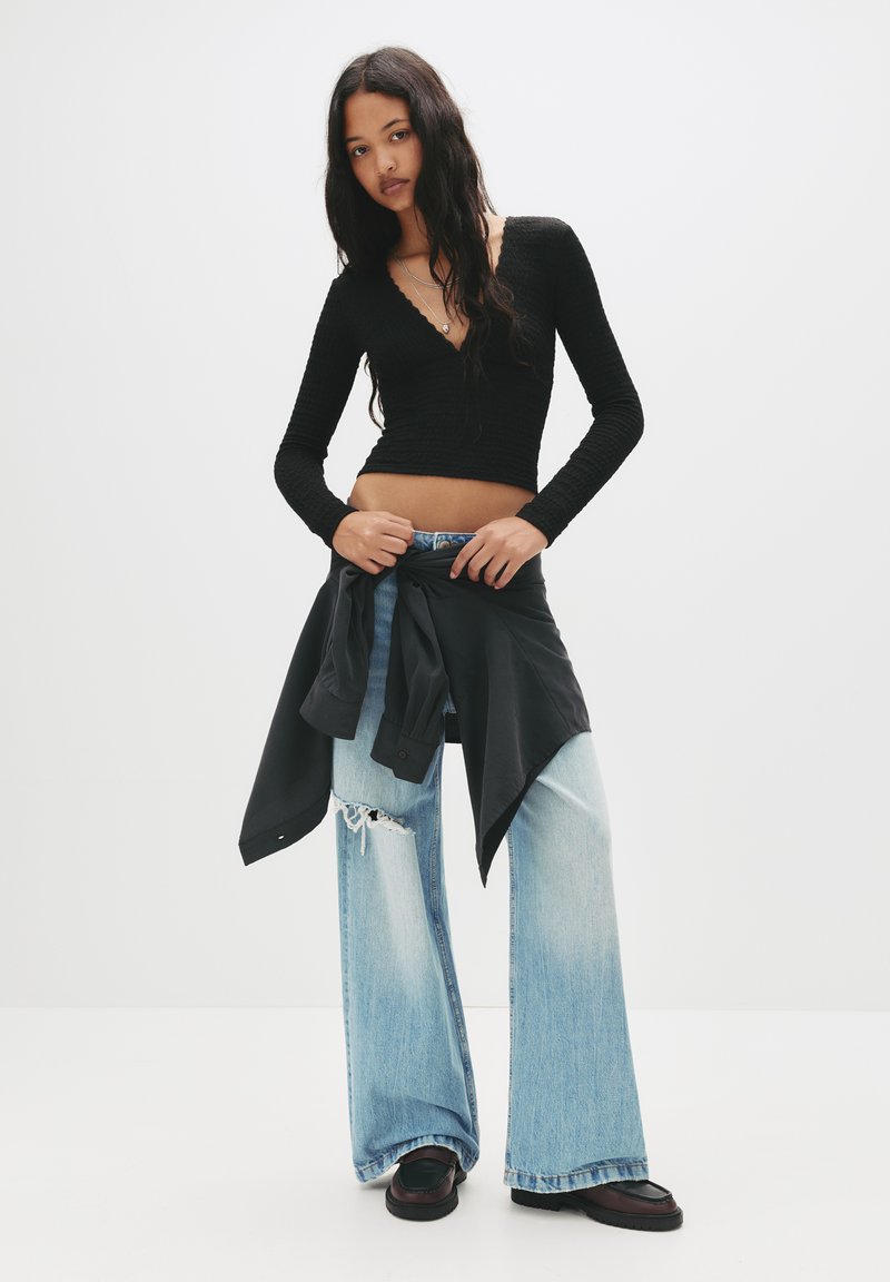 Schwarz gestreifte Langarmbluse mit tiefem V-Ausschnitt, kombiniert mit hellblauen, weit geschnittenen, distressed Jeans. Eine schwarze Jacke ist um die Taille gebunden.