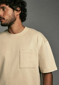 Beige bomulds T-shirt med rund hals og korte ærmer. Har en rektangulær brystlomme med dobbelt søm. Glat tekstur.