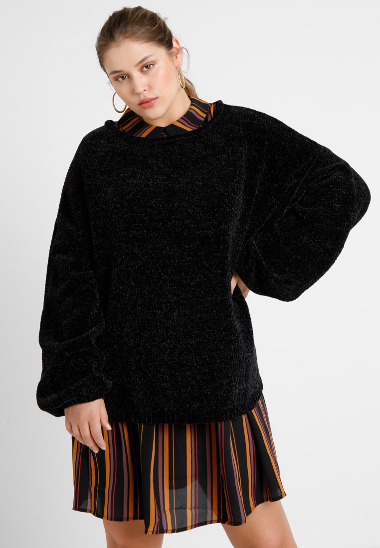 Black fuzzy sweater with oversized sleeves, layered over a multicolored striped dress featuring orange, yellow, and black lines.
