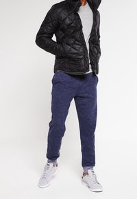 Schwarze gesteppte Jacke, graues T-Shirt, blaue Jogginghose und weiße Sneakers. Die Jacke hat einen hohen Kragen, einen Reißverschluss und Seitentaschen.