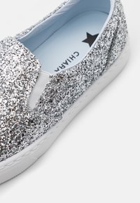 Hopeiset kimalteet slip-on kengät valkoisella kumipohjalla, joustavat sivupaneelit ja vaaleansininen sisäpohja, jossa on musta tähti-logo.