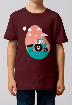 Kind trägt ein kastanienbraunes T-Shirt mit einem blauen Traktor, der einen Anhänger zieht, vor einem rosa ovalen Hintergrund mit weißen Wolken.