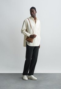 Svijetla košulja dugih rukava, tamne ravne hlače, kremasti tenisice i mala smeđa torbica preko ramena. Jednostavan dizajn i neutralne boje.