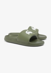Green rubber slide sandals with a wide strap featuring a white crocodile logo, textured footbed, and a flat sole. Lacoste branding on the side.