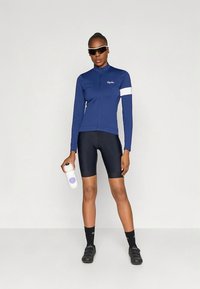 Tumesinine rattasõidu jope valge aktsendiga, mustad lühikesed püksid ja valge joogipudel. Päikeseprillid ja spordijalatsid täiustavad sportlikku välimust.