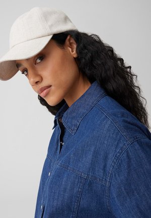 Giovane con lunghi capelli ricci che indossa un berretto beige e una camicia di jeans blu con bottoni, di profilo leggermente laterale su uno sfondo semplice.