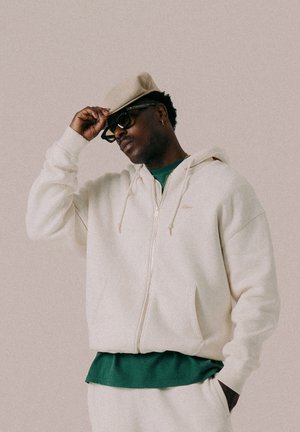 Cremefarbener Zip-Hoodie aus weichem Stoff, mit einer grünen Unterlage und einer stilvollen Flachkappe. Der Träger trägt dunkle Sonnenbrillen und hat strukturierte Haare.