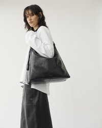 Donna con capelli scuri che indossa una maglietta bianca e una gonna di pelle nera, mentre porta una grande borsa a spalla in pelle nera su uno sfondo semplice.