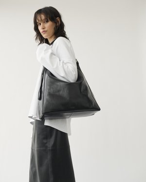 Femme aux cheveux foncés portant un haut blanc et une jupe en cuir noir, portant un grand sac à bandoulière en cuir noir sur un fond uni.