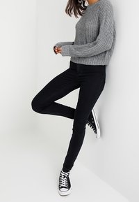 Gray knitted sweater with a crew neckline, paired with black skinny jeans and black high-top sneakers. The outfit leans against a white wall.