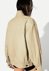 Beige Overshirt-Jacke mit lockerer Passform, hochgeschlossenem Kragen, geripptem Saum und schwarzen Knopfakzenten. Sanfte Textur mit minimalen Nähten.