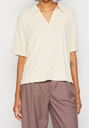 Personne portant une chemise crème à manches courtes avec col en V et un pantalon ample mauve, montrant un tatouage géométrique sur l'avant-bras gauche.