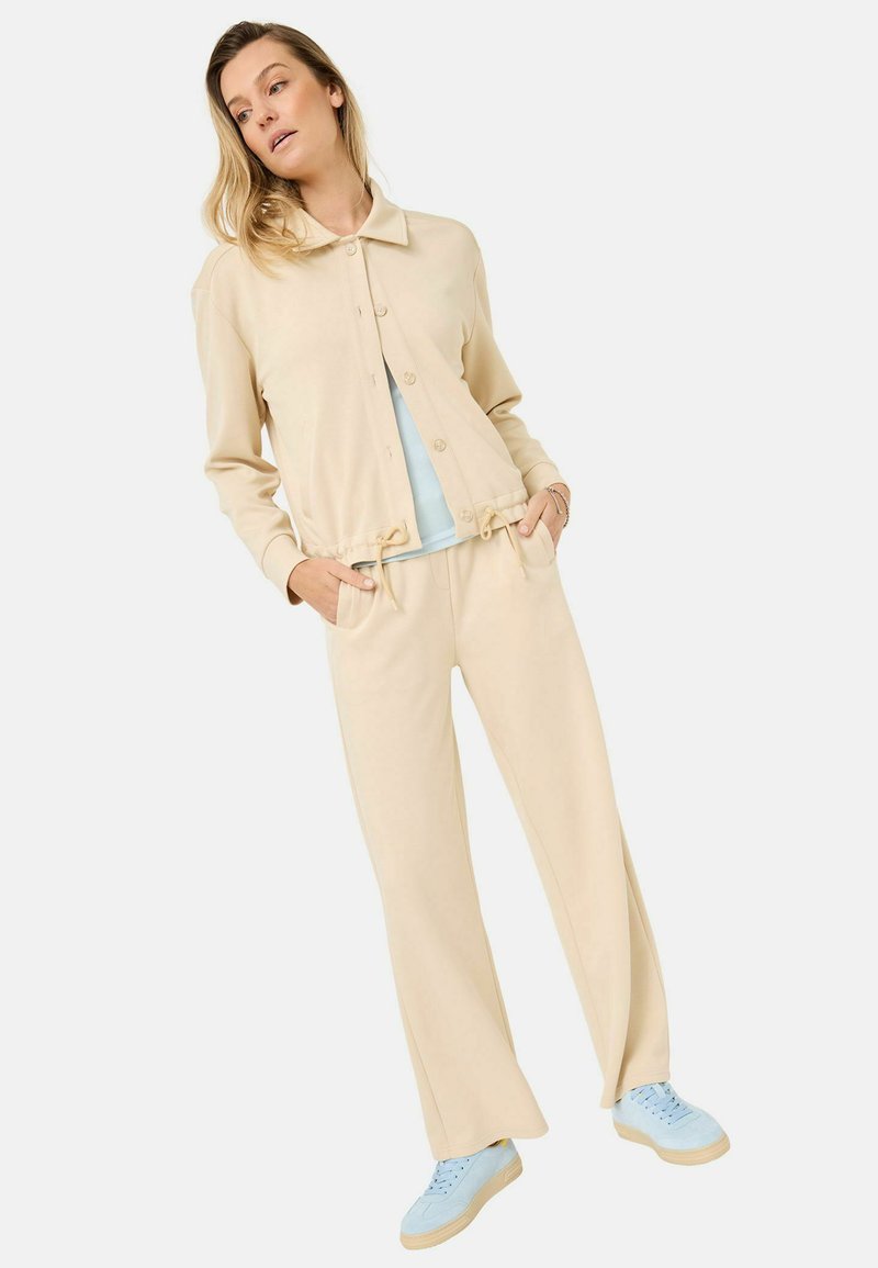 Completo coordinato beige chiaro composto da una giacca a bottoni cropped e pantaloni a gamba larga, entrambi in tessuto morbido. Abbinato a scarpe da ginnastica blu chiaro.