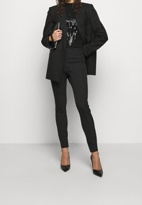 Chaqueta de traje negra sobre un top negro con lentejuelas, combinada con pantalones negros ajustados y tacones de punta negra. Tejido suave y diseño ajustado.