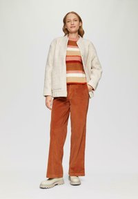 Helle graue Wollmischjacke, rostfarbene Cordhosen und ein gestreifter Strickpullover in warmen Farben. Klobige beigefarbene Stiefeletten runden das Outfit ab.