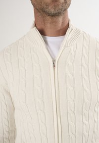 Cardigan crema a coste con colletto alto, zip frontale e design testurizzato. La morbida lana spessa crea un aspetto accogliente.