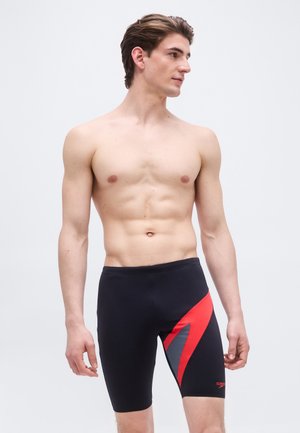 Muskulöser Mann trägt schwarze knielange Badeshorts mit roten und grauen eckigen Mustern, schaut nach rechts vor einem schlichten Hintergrund.