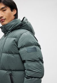 Tumman vihreä puhvijakku, jossa on pehmustettu muotoilu, hupullinen, vetoketjulliset taskut ja Marc O'Polo -logotunnus hihassa.