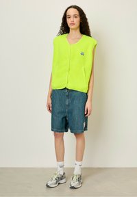 Neon green fleece vest with a zipper, featuring two side pockets. Paired with denim shorts and gray sneakers with accents.