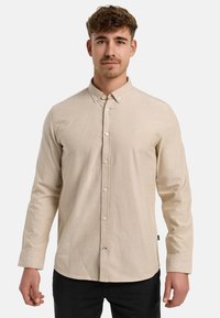 Camisa de manga larga beige con cuello abotonado, cierre frontal de botones y un pequeño logo en el pecho. Textura de tejido suave.