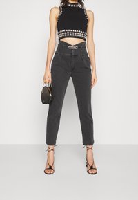 Top corto negro con tachuelas, pantalones oscuros de tiro alto de color gris, y tacones negros con tiras. La modelo sostiene un pequeño bolso negro.