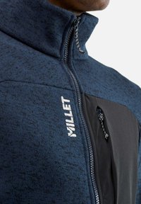 Veste en polaire bleu foncé avec une surface texturée, panneau noir, col haut et poche poitrine zippée. Dotée d'un logo blanc.
