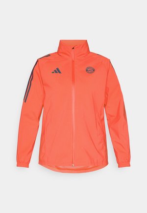Klart orange zip-up jacka med hög krage. Har mörkblå ränder på axlarna och en rund logotyp på bröstet. Slät textur.