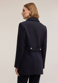 Abrigo azul marino de mezcla de lana con una silueta entallada, que presenta un cuello con muescas, un detalle de botón en la parte posterior y una tela texturizada.