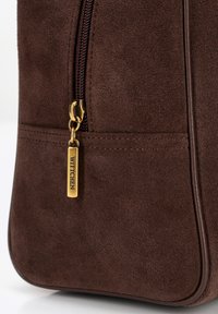 Bolso de ante marrón con un tirador de cremallera dorado y una etiqueta rectangular. Textura suave con sutiles detalles de costura.