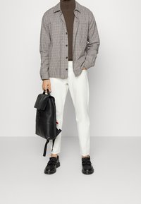Tenue masculine comprenant une veste à carreaux marron et blanche, un col roulé marron, un pantalon blanc, des chaussures noires et un sac à dos noir avec un rabat.