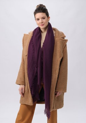 Cappotto in finta pelliccia beige, design oversized, con tasche frontali. Sciarpa viola profondo con bordi frangiati. Modella indossa un maglione beige e pantaloni marroni.