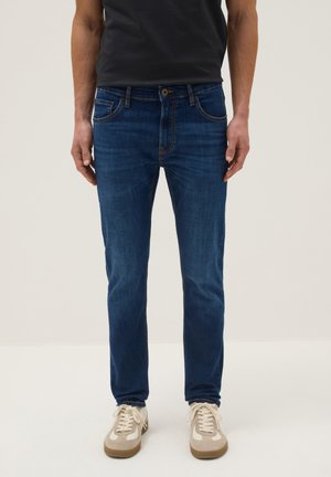 Man draagt een slim-fit donkerblauwe spijkerbroek, witte en beige sneakers en een zwart T-shirt, staand tegen een effen lichte achtergrond.