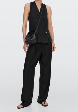 Žena nosící černý bezrukávový blejzr, široké černé kalhoty, černou pletenou kabelku přes rameno a černé žabky stojící před bílým pozadím.