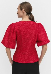 Kobieta z brązowymi włosami związanymi z tyłu, ubrana w czerwoną bluzkę z bufkami o fakturze i czarne spodnie, widoczna od tyłu na jednolitym tle.