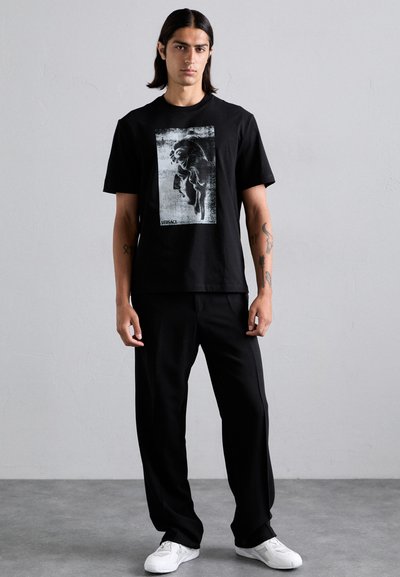 T-shirt en coton noir avec un grand imprimé graphique blanc, associé à un pantalon noir ample et des baskets blanches.