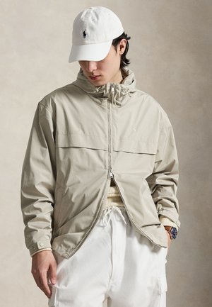 Mand iført beige hættejakke, hvid striktrøje, hvide cargobukser, hvid kasket og armbåndsur, står foran en neutral baggrund.