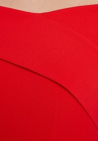 Tissu texturé rouge avec un décolleté en biais. Le design présente une forme lisse et ajustée avec des lignes épurées, sans ornements.