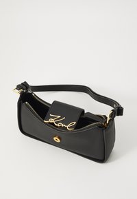 Black leather handbag with gold hardware, curved design, front zip pocket, and a flap featuring the word "Karl" in script. Adjustable strap.