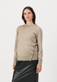 Beige neulottu neulepaita pyöreällä kaula-aukolla, ribbireunoilla ja helmassa, jossa on nappiyksityiskohta vasemmalla puolella. Mallilla on päällään mustat hame.