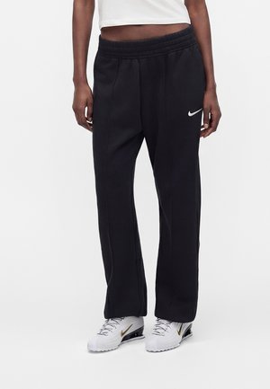 Osoba ubrana w luźne czarne spodnie dresowe Nike z białym logo, biały krótki top oraz białe sneakersy Nike ze złotymi łyżwami, stojąca wyprostowana.