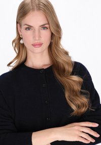 Cardigan blu navy con bottoni realizzato in morbido tessuto a maglia, con scollo rotondo e polsini a costine. Capelli acconciati in onde morbide.