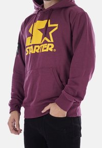 Sudadera granate hecha de un tejido de mezcla de algodón que presenta un gran logo amarillo con una "S" y una estrella. Tiene un bolsillo tipo canguro y puños acanalados.