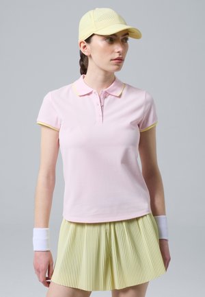 Donna che indossa una polo rosa pallido, gonna gialla plissettata, cappellino giallo e polsini bianchi, in piedi davanti a uno sfondo grigio uniforme.