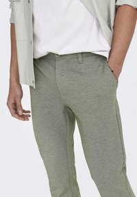 Pantalones de color verde claro, texturizados y elásticos con un ajuste tapered, que cuentan con un cierre frontal con botones y bolsillos laterales. Llevados con una camisa ligera.