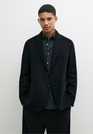 Ung mand med kort hår iført en sort blazer over en mørk knap-skjorte, stående med hænderne i lommerne mod en ensfarvet lys baggrund.