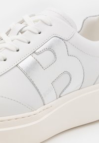 Bílé kožené tenisky s metalickým stříbrným logem "B", texturovanými detaily, kulatými tkaničkami a krémovou gumovou podrážkou.