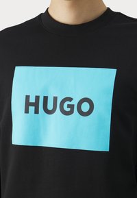 Svart sweatshirt av bomull, med en stor ljusblå rektangulär grafik med ordet "HUGO" i fet svart font i mitten.