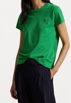 Frau trägt ein hellgrünes T-Shirt mit kurzen Ärmeln und einem kleinen gestickten blauen Pferdelogo auf der Brust, kombiniert mit dunkler Hose.